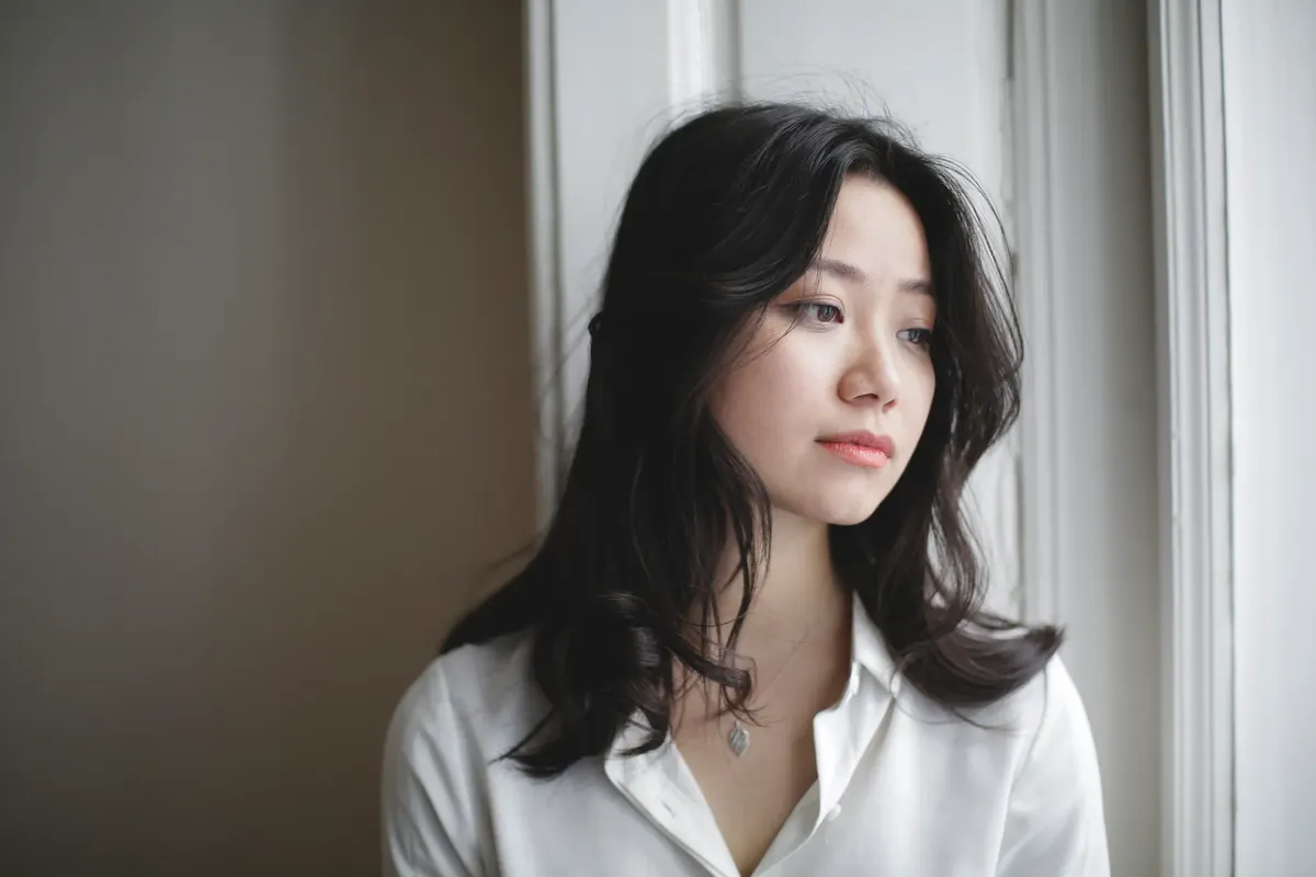 Young Asian female looking out window and dreaming while resting at home on weekend day