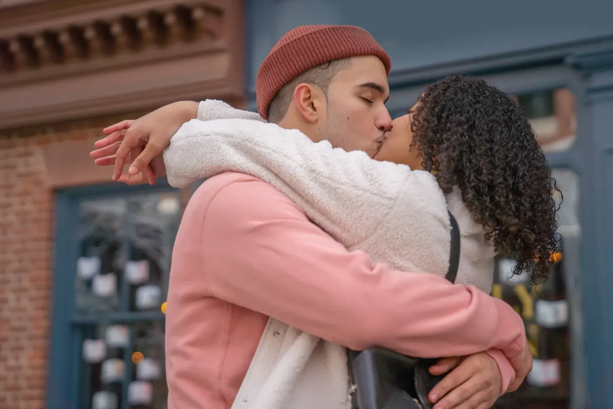 Side view of happy romantic ethnic couple with eyes closed kissing each other on blurred background of urban building