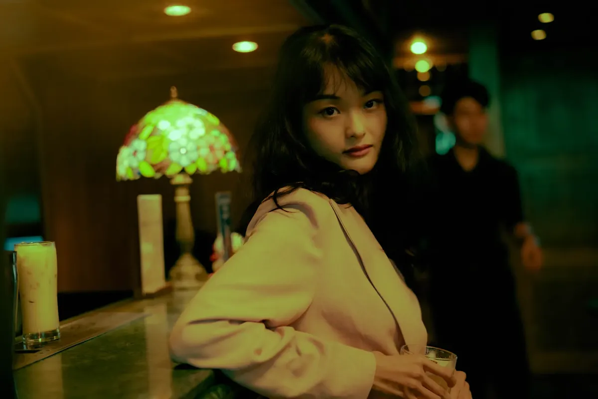 Stylish Asian woman in suit jacket posing by the bar in a cozy, warmly-lit setting. Fashionable evening moment.