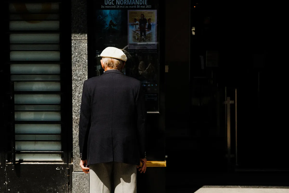 Elderly man in classic attire viewing movie posters outside a cinema.
