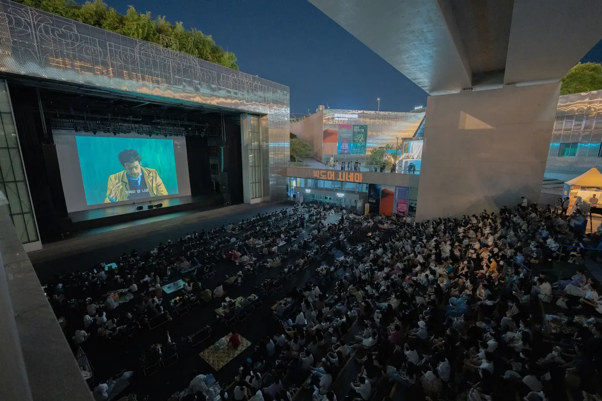 A large audience gathers outdoors at night to watch a movie screening under a bridge.