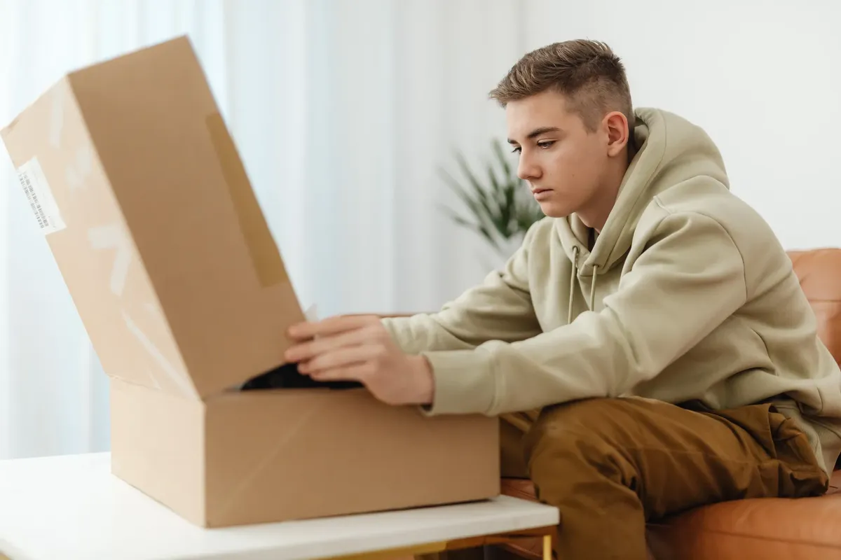 Teenage boy unboxing a large cardboard package while sitting on a couch in a warm indoor setting.