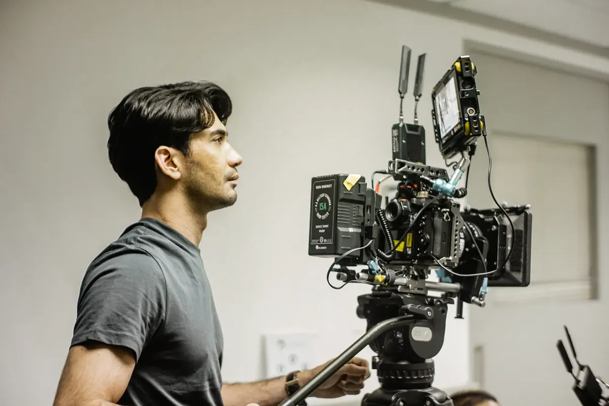 A professional videographer in a gray shirt operates a high-end camera setup indoors, capturing footage in a studio environment.
