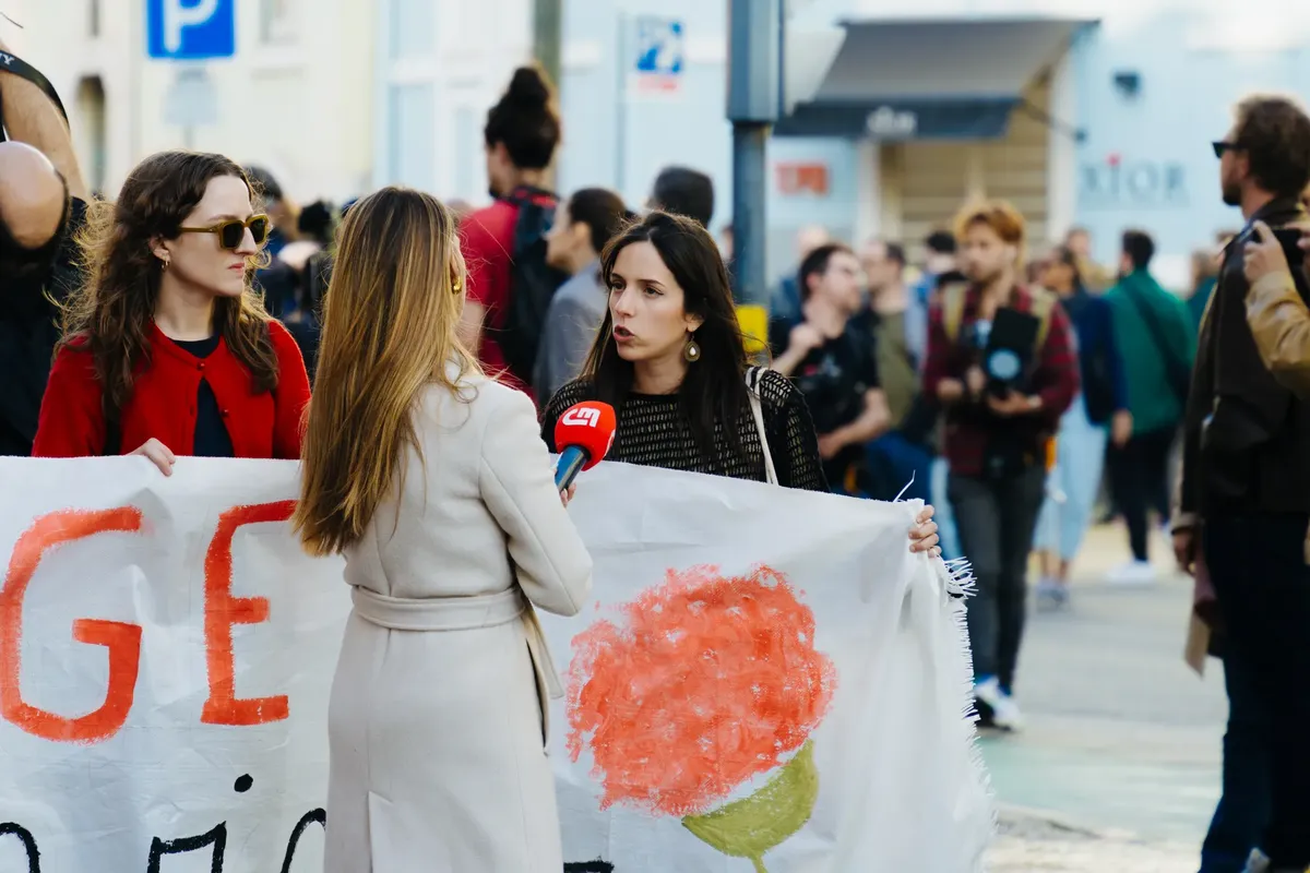 Journalist interviews protesters during a city rally with people holding banners and flags.