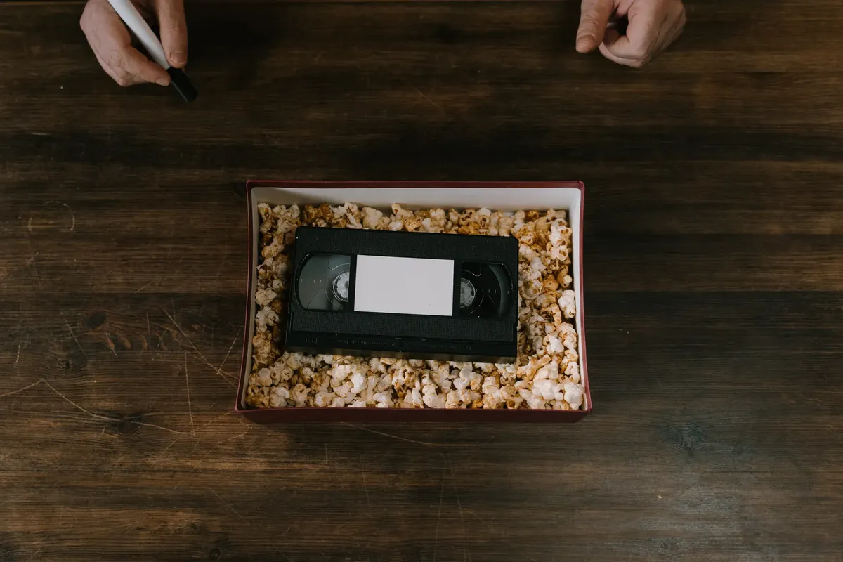 A VHS tape surrounded by popcorn in a box, placed on a wooden surface. Hands holding a marker are also visible.
