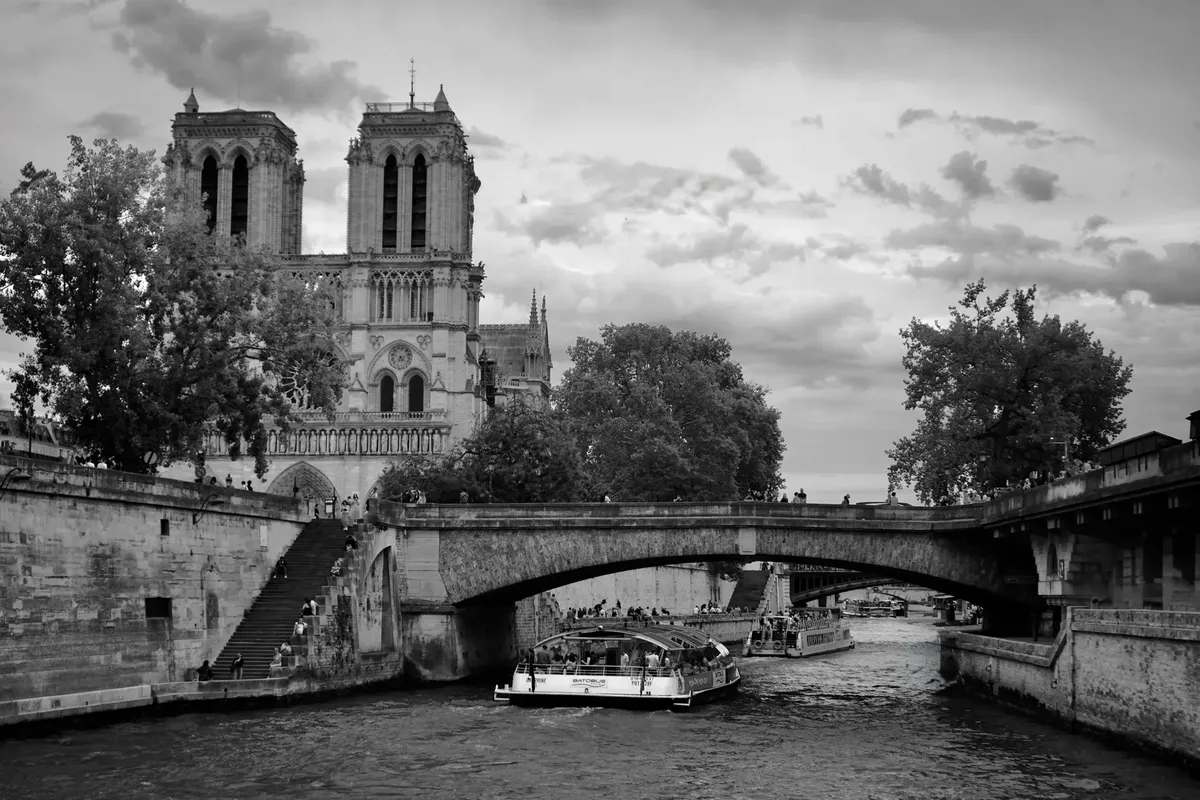 Black and white photo of Notre-Dame Cathedral and the Seine River in Paris with boat and bridge.
