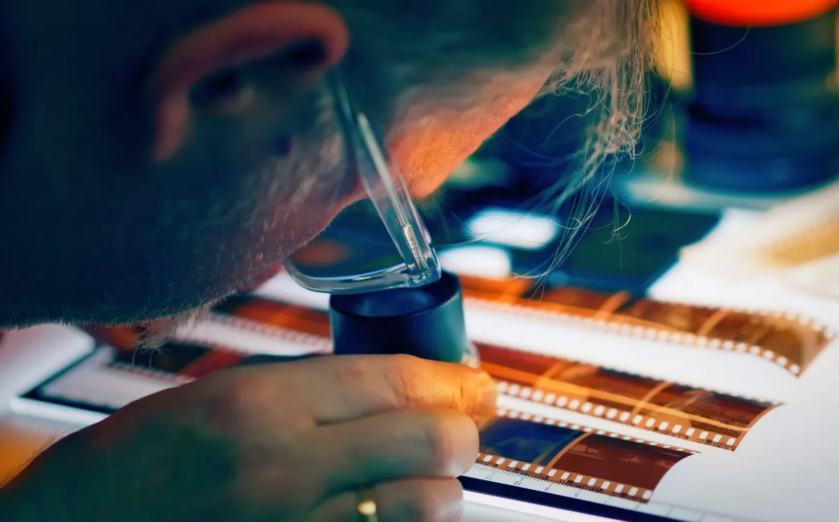A photographer closely inspects film negatives using a loupe and lightbox in a creative studio setting.