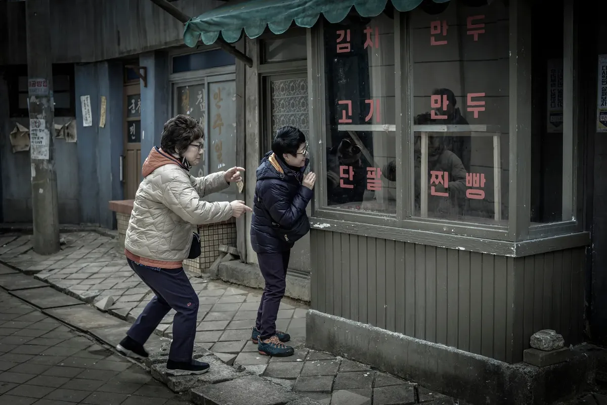 Two elderly women peering through a shop window with Korean signs in an urban setting.