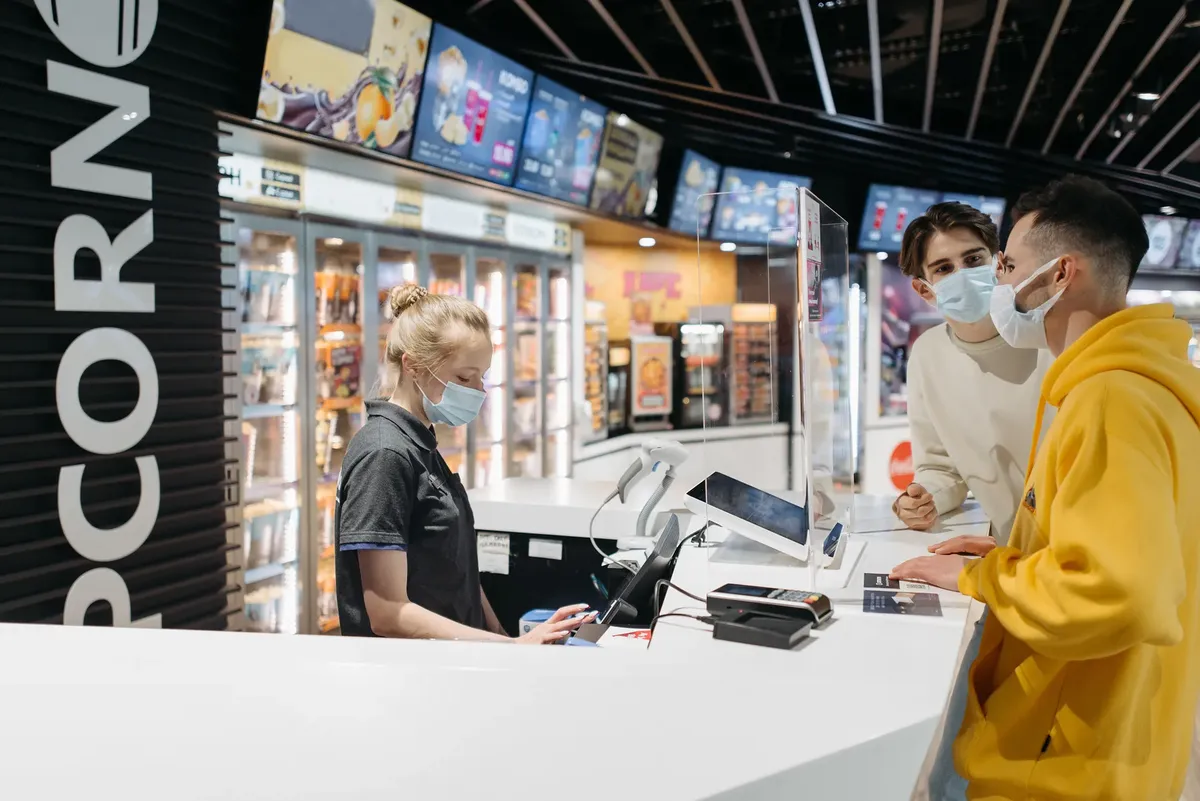 Customers wearing masks interact with a cashier at a cinema concession stand, following safety protocols.