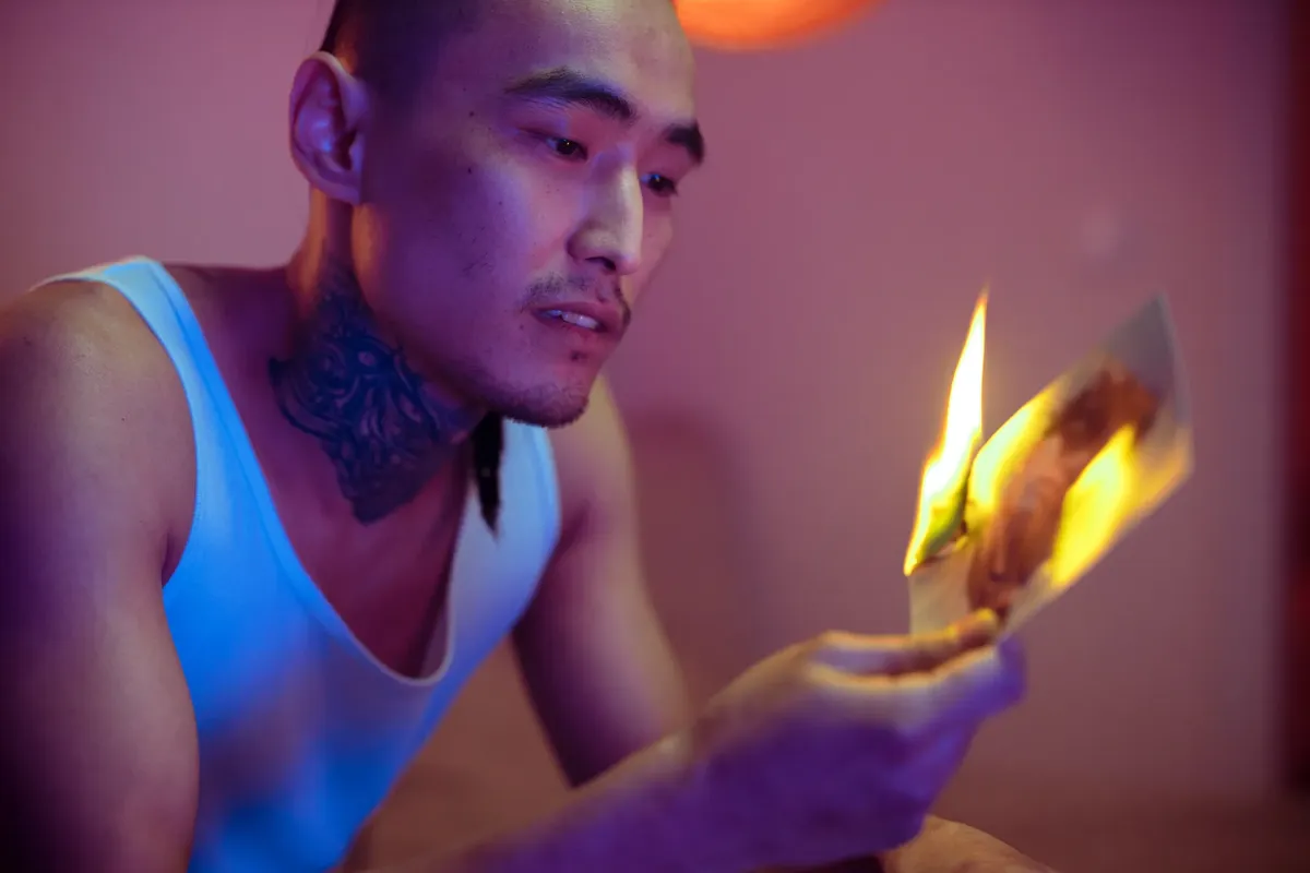 A tattooed man in a tank top burning a photograph indoors. Moody and dramatic lighting.