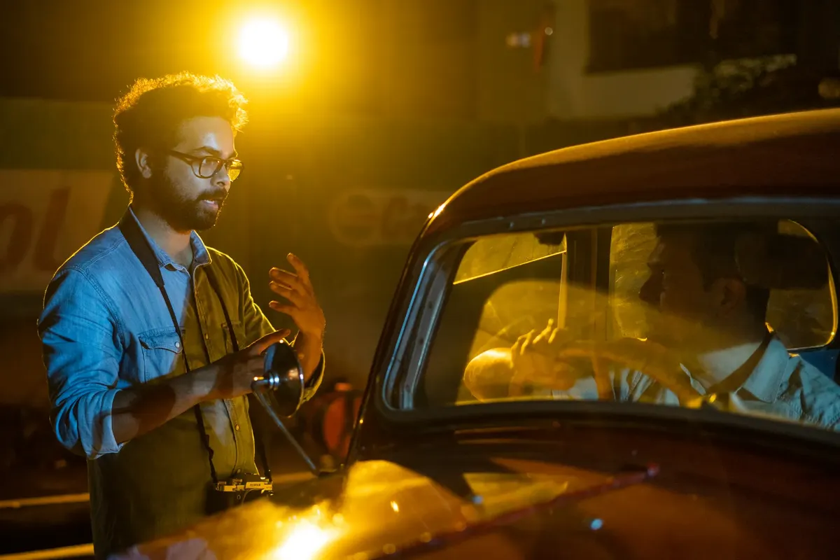 Director and actor on a vintage car movie set at night under bright lighting.