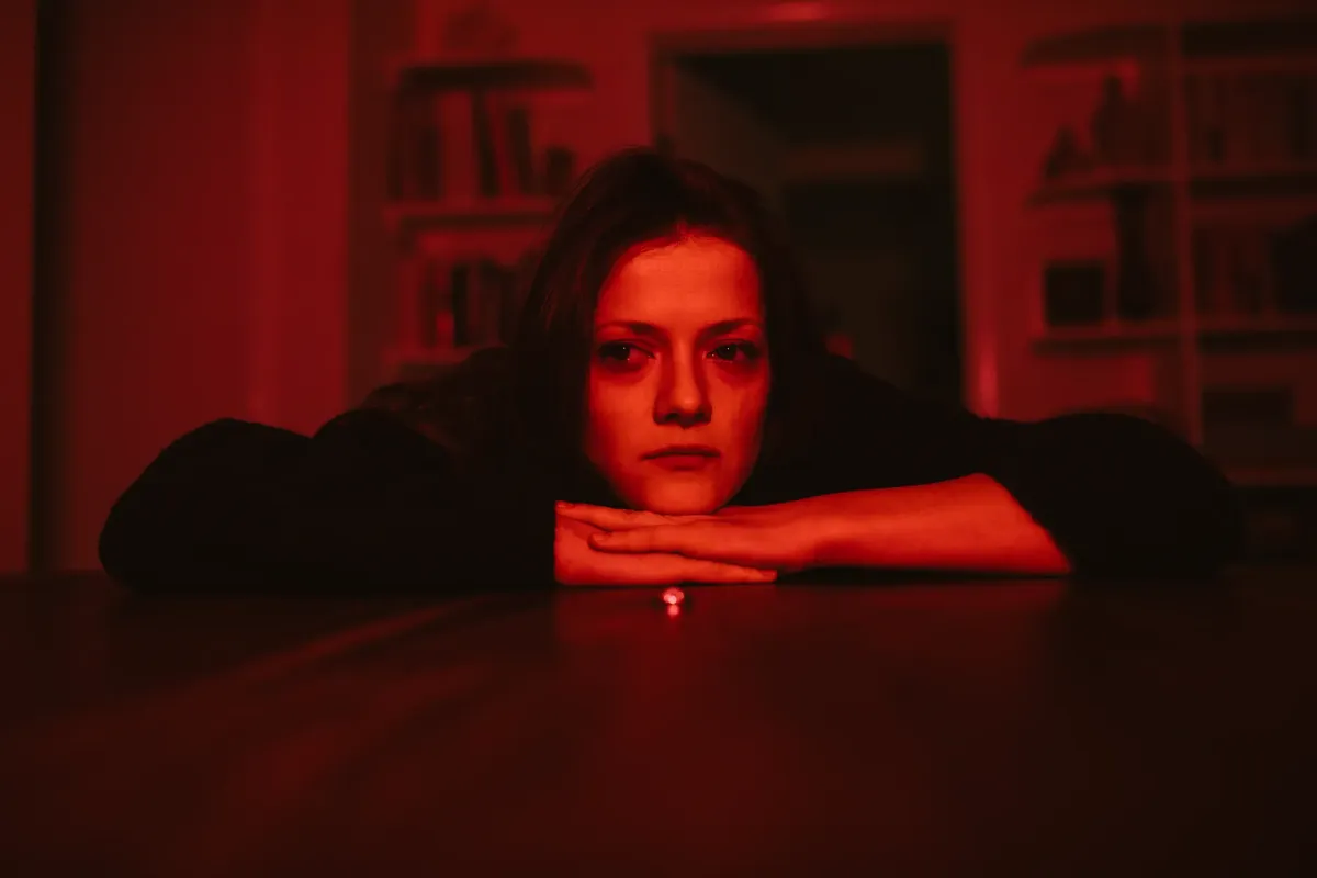 Young woman sitting at a table looking pensive, illuminated by red light, creating a dramatic atmosphere.